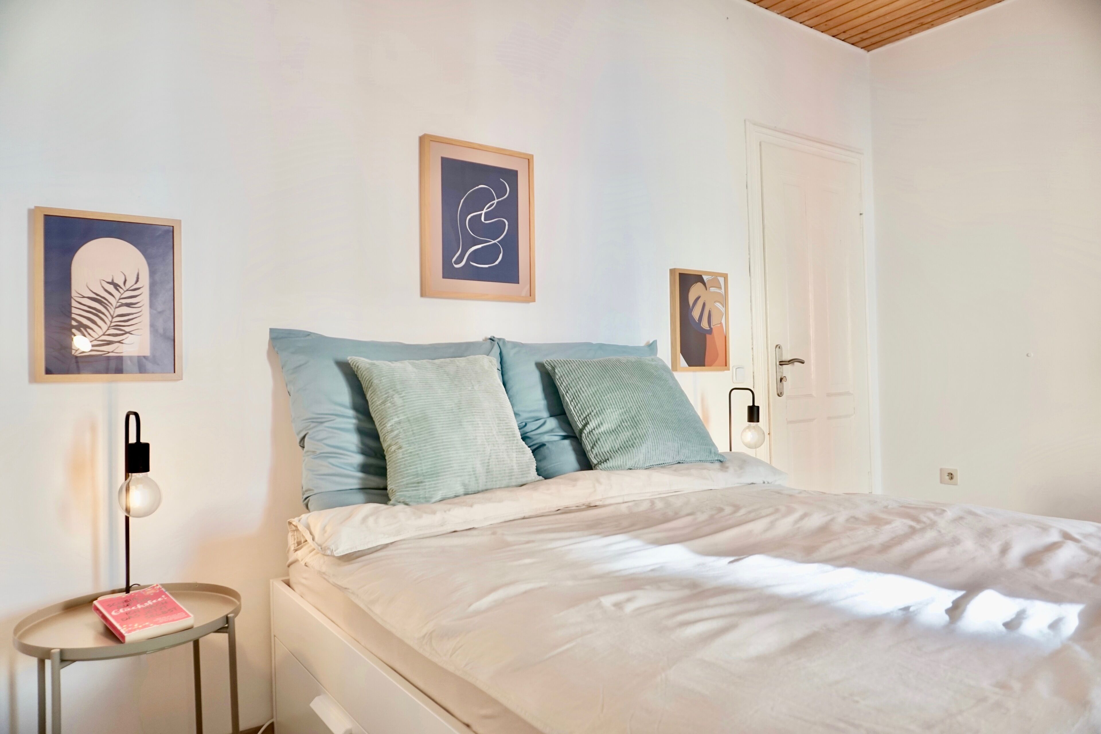 Une chambre à coucher avec un lit, des oreillers, une table de chevet avec une lampe et des œuvres d’art encadrées accrochées aux murs.