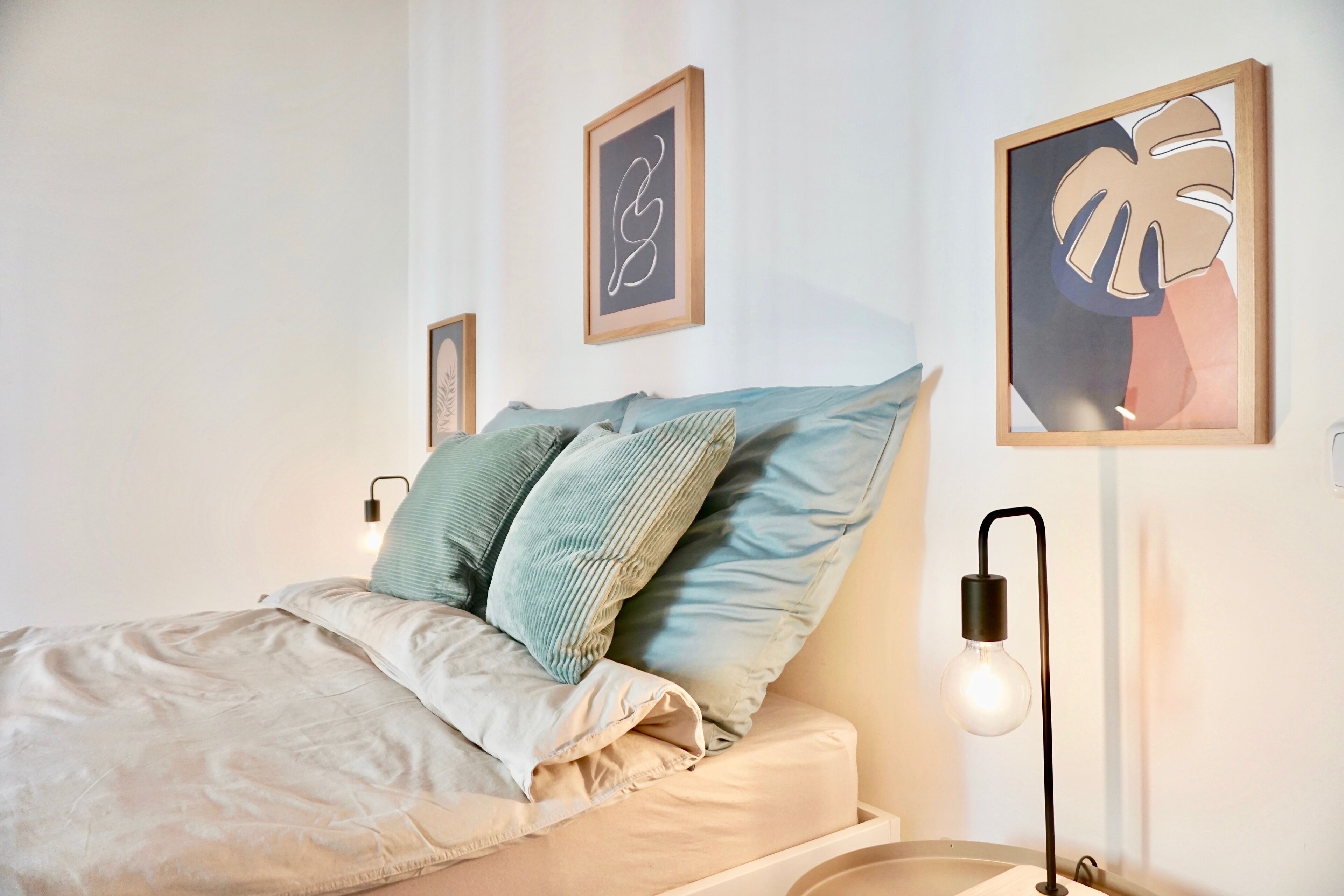 Una cama bien hecha con un edredón de color claro y dos almohadas, una mesita de noche con una lámpara, y dos obras de arte enmarcadas en la pared.