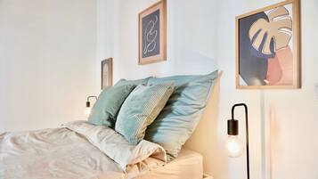 Una cama bien hecha con un edredón de color claro y dos almohadas, una mesita de noche con una lámpara, y dos obras de arte enmarcadas en la pared.