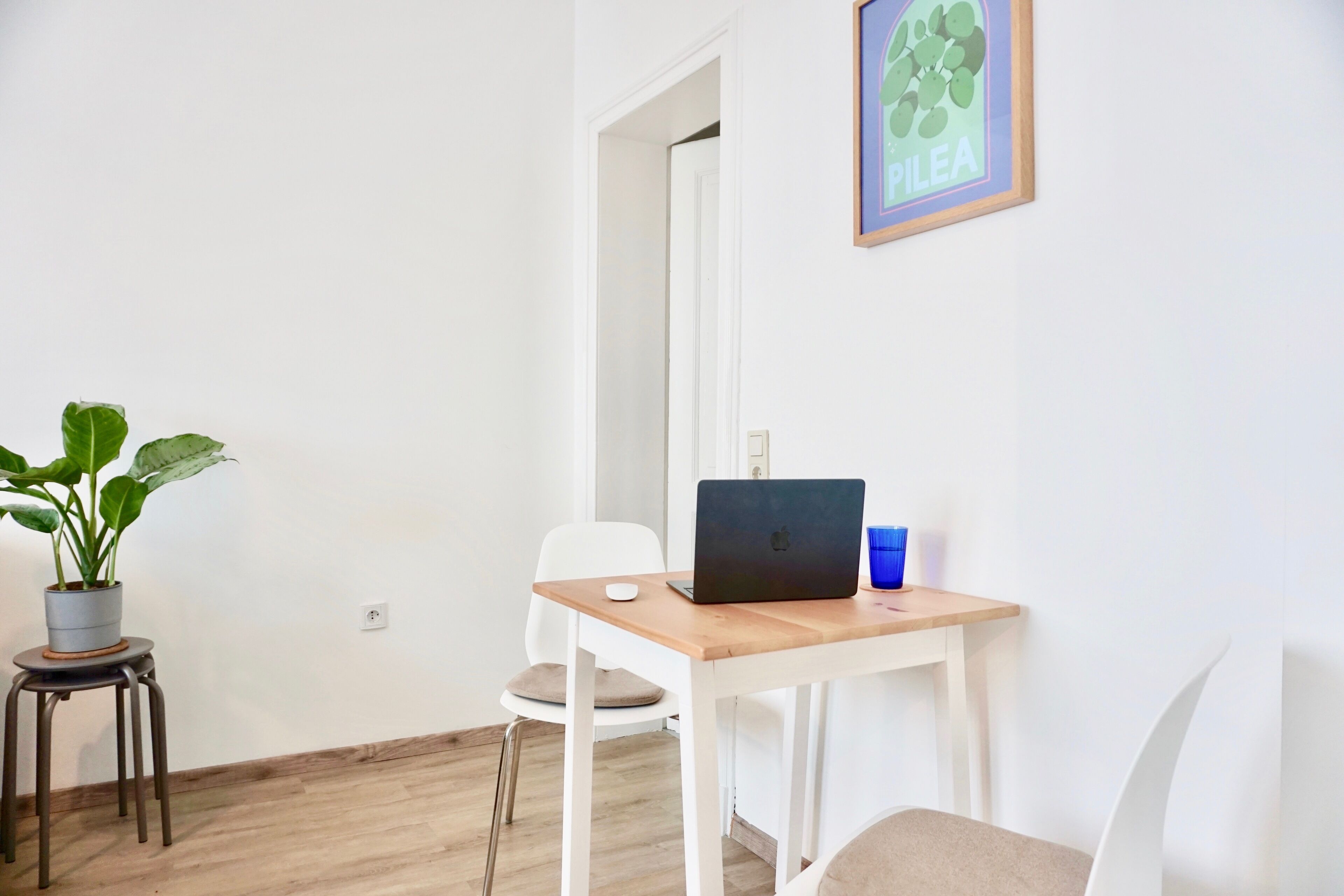 A small, clean room with a wooden table, a laptop, a potted plant, and a framed picture on the wall.