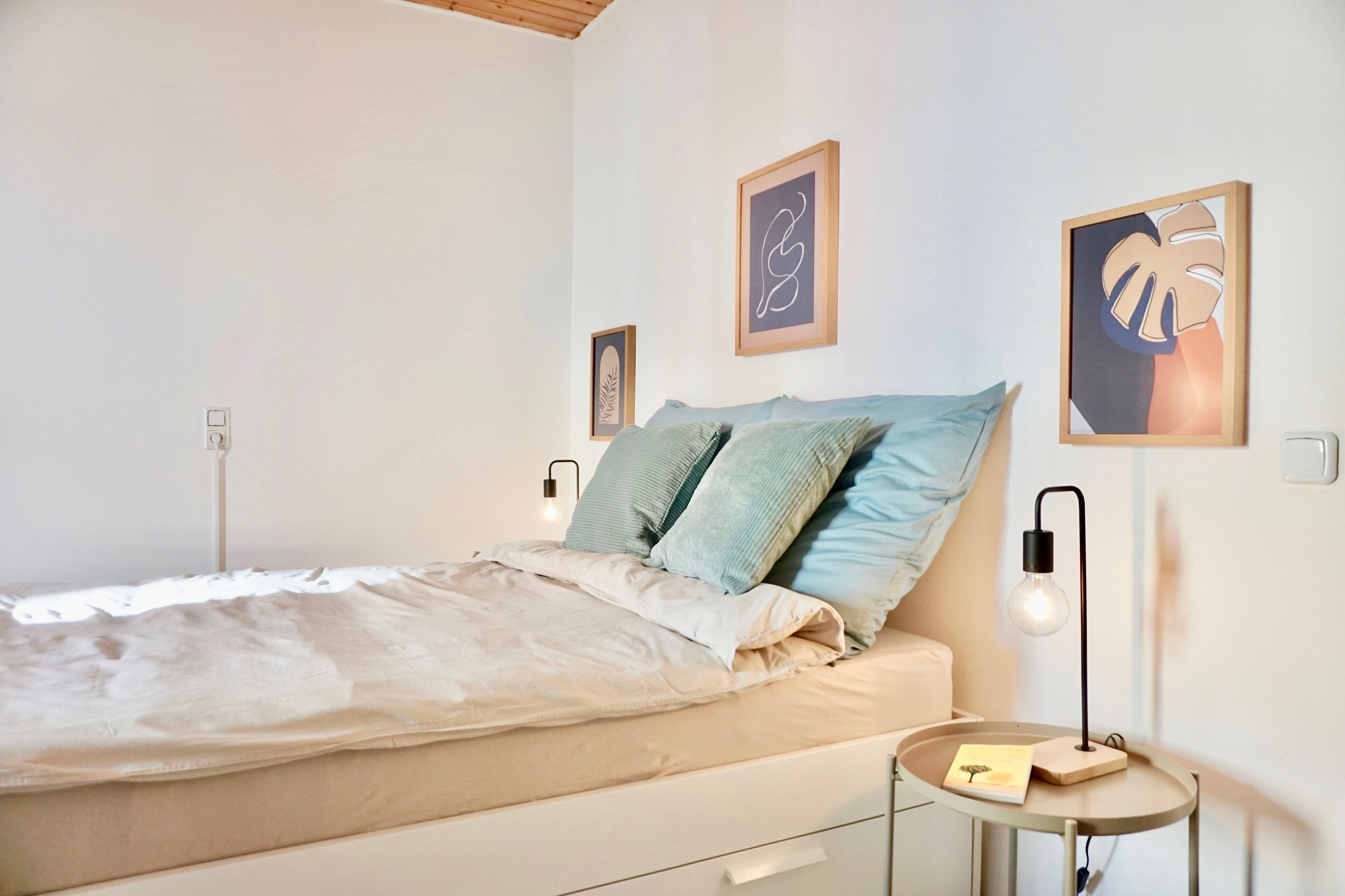 A neatly made bed with a light-colored comforter and two pillows, a bedside table with a lamp, and framed artwork on the wall.