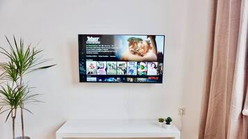 A flat-screen TV mounted on a wall above a white TV stand with a small plant and a book on it.