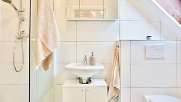 Un baño moderno con cabina de ducha de vidrio, un lavamanos blanco con espejo y un inodoro con un dispensador de papel higiénico visible.