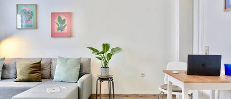 Ein modernes Wohnzimmer mit einem grauen Sofa, einem kleinen Holztisch mit einem Laptop, einer Topfpflanze und zwei gerahmten Bildern an der Wand.