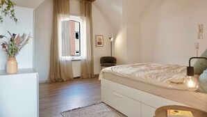 Een slaapkamer met een houten plafond, een bed met een witte matras, een bedlampje, een klein tafeltje en een vaas met gedroogde bloemen.