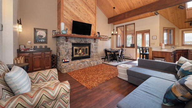A spacious living room with a stone fireplace, wooden ceiling beams, a television, and a dining area with a kitchen in the background. - Kings Crossing Townhome 12 New Downtown 3 bedroom Private Hot Tub (Winter Park)