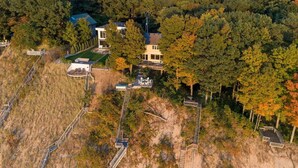 An aerial view of a property with multiple buildings, including a main house and several smaller structures, surrounded by dense forest and a sandy area with wooden walkways.