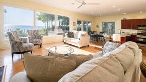 A spacious living room with a large sofa, armchairs, and a glass coffee table. There is a kitchen area with a dining table and chairs, and a view of the ocean through large windows.