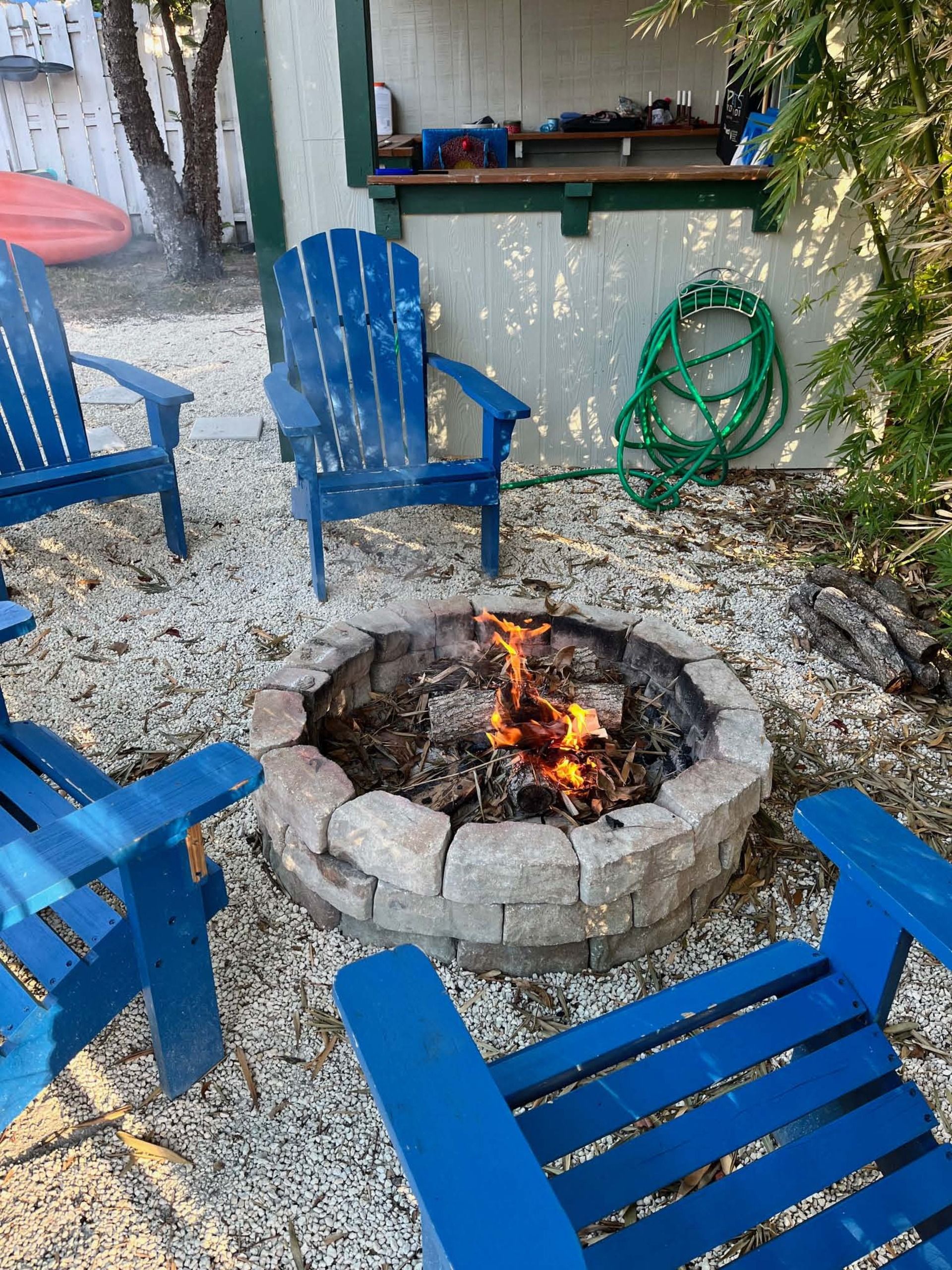 A fire pit surrounded by blue Adirondack chairs, a green garden hose, and a wooden deck.