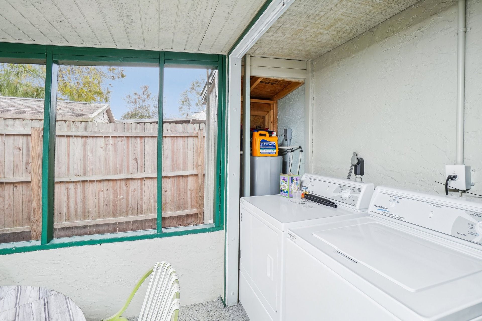 A laundry room with a large white washer and dryer, a wooden fence outside, and a green-framed window.
