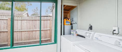 A laundry room with a large white washer and dryer, a wooden fence outside, and a green-framed window.