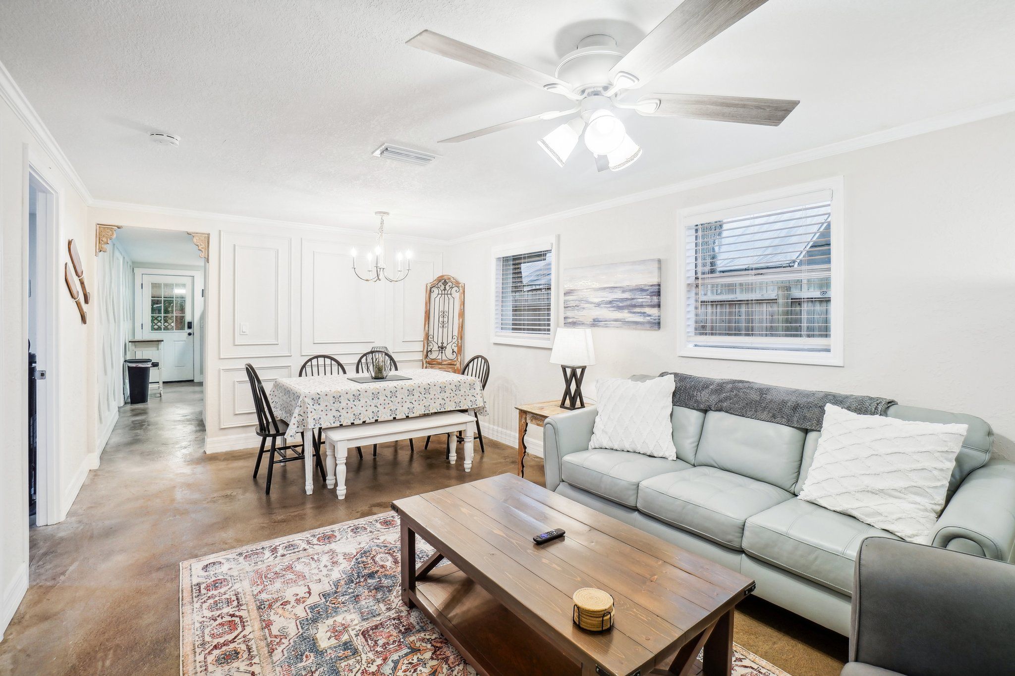A spacious living room with a ceiling fan, a dining area with a table and chairs, a sofa with cushions, and a large window with blinds.