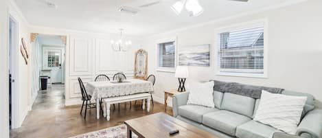 A spacious living room with a ceiling fan, a dining area with a table and chairs, a sofa with cushions, and a large window with blinds.