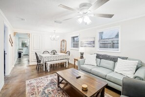 A spacious living room with a ceiling fan, a dining area with a table and chairs, a sofa with cushions, and a large window with blinds. - 3BR Riverside Escape ~ Hot Tub, Clear Kayaks & BBQ (Spring Hill)