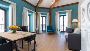 A room with a wooden dining table, chairs, a sofa, and a blue armchair. There are curtains on the windows, a lamp on a side table, and a vase with flowers on the table. - Florella Residences 2 pièces Antibes (Cannes)