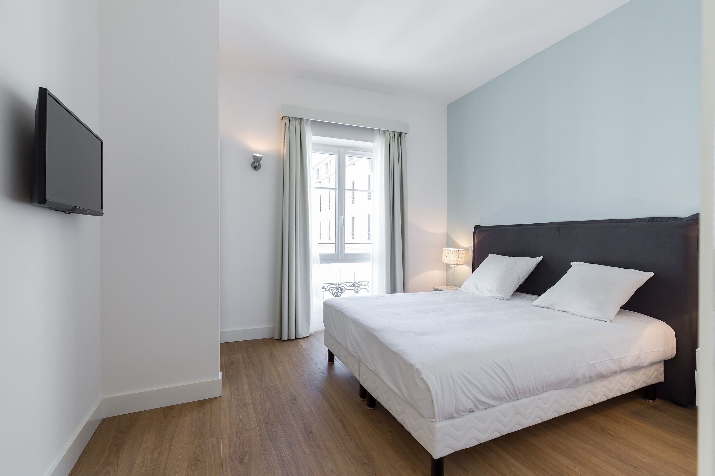 Une chambre à coucher comprenant un lit, un téléviseur à écran plat fixé au mur, une fenêtre avec des rideaux et une table de chevet avec une lampe.