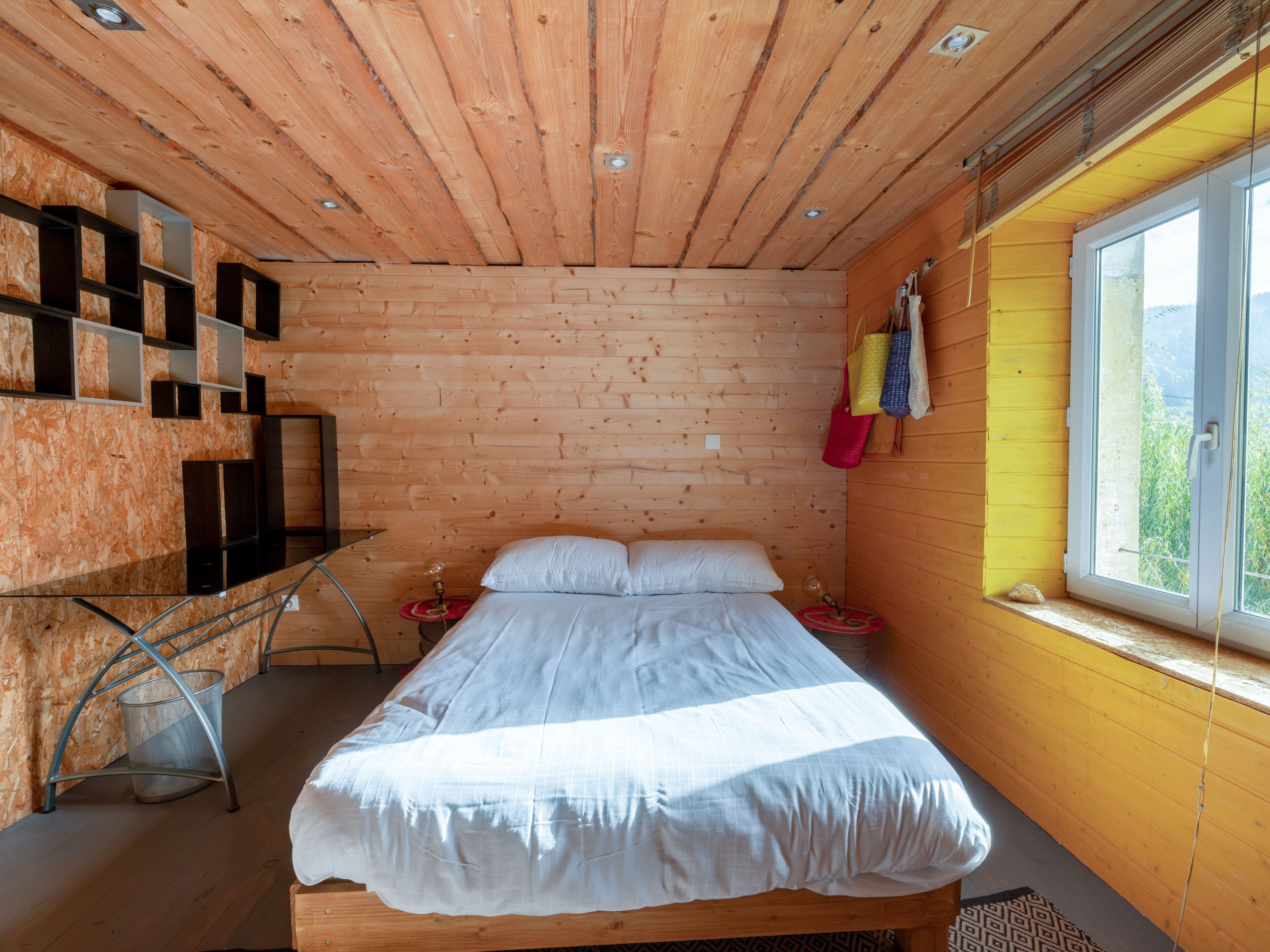 A wooden cabin bedroom with a bed, a small desk, and a window with a view.