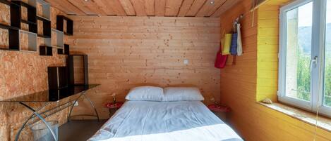 A wooden cabin bedroom with a bed, a small desk, and a window with a view.