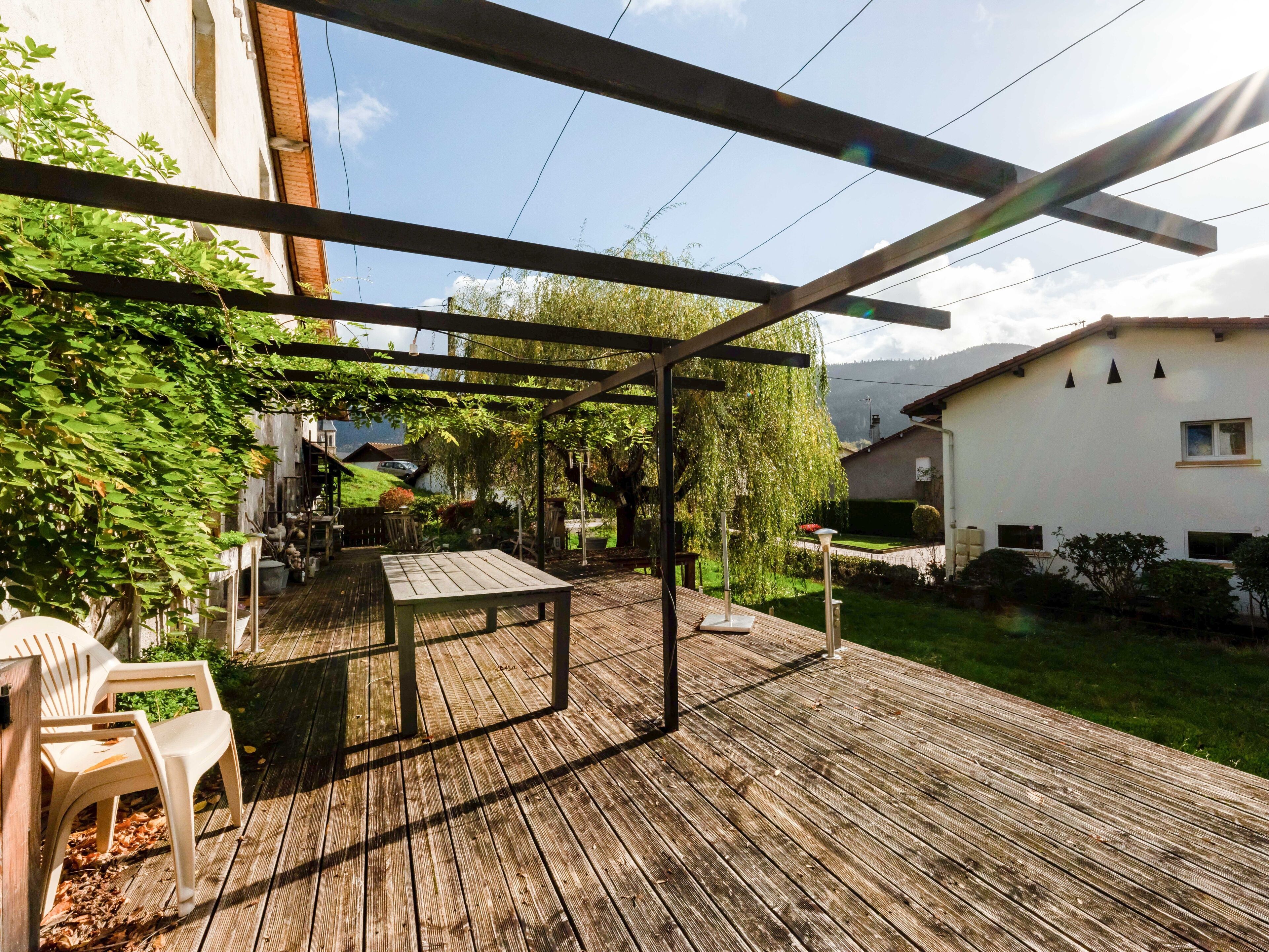 A wooden deck with a pergola, a table, and chairs, surrounded by greenery and a white building.