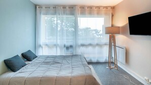 Une chambre à coucher avec un lit, une lampe sur pied, un téléviseur et une fenêtre avec des rideaux légers.