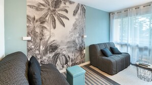 Un salon moderne avec deux canapés à motifs, une grande fresque murale représentant un feuillage tropical, une petite table ronde et des rideaux légers à une fenêtre.