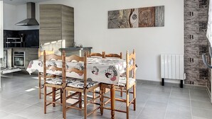 Un comedor con una mesa preparada para cuatro personas, rodeada de sillas de madera con cojines estampados. Una lámpara colgante moderna ilumina el espacio, y un gran cuadro abstracto decora la pared.