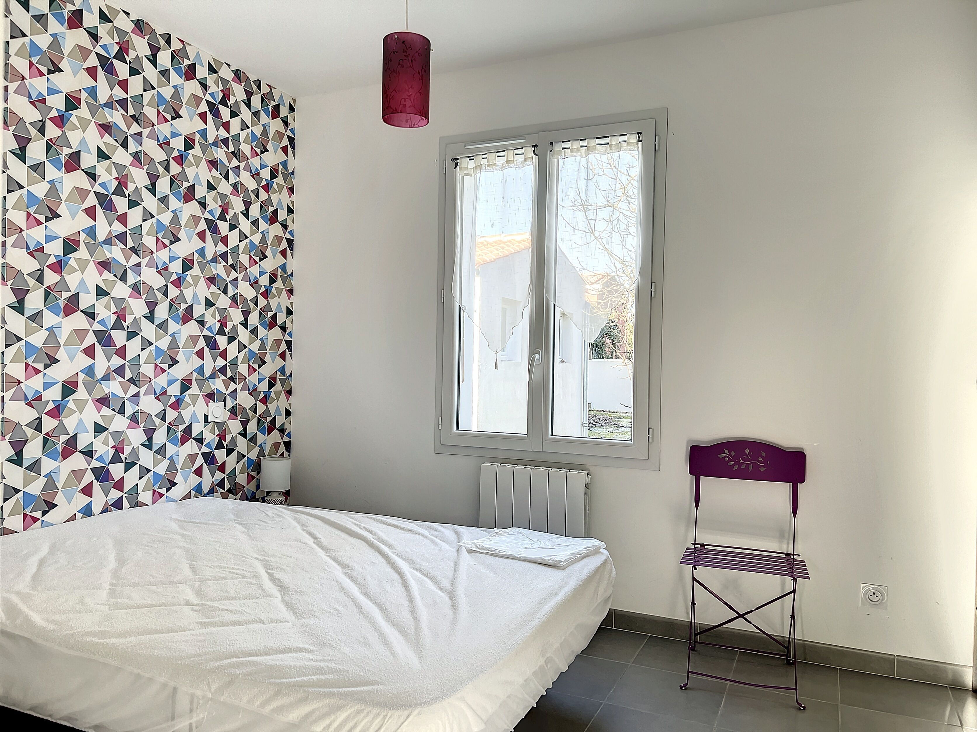 A bedroom with a bed, a radiator, a window with curtains, and a wall with a geometric pattern.