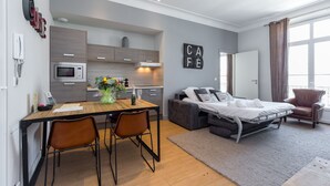 A modern hotel room with a kitchenette, a bed with a grey headboard, a brown leather armchair, and a large window with curtains.
