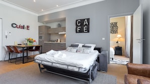 A modern bedroom with a bed, a desk, and a kitchenette in the background.