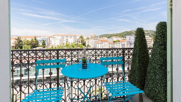 A balcony with a blue table and chairs, a bottle of wine, and a view of buildings and trees.