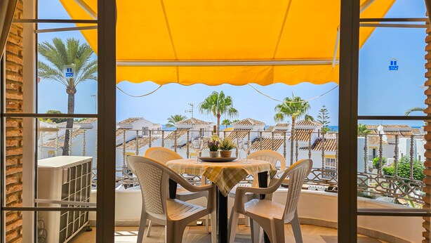 Un balcón con toldo amarillo, sillas de mimbre y una mesa con un florero y copas de vino.