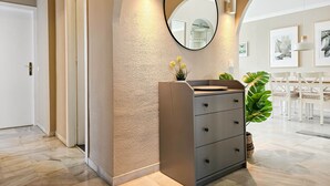 Un pasillo con pared texturizada, un espejo redondo, un aparador gris y un comedor con sillas blancas y una mesa.