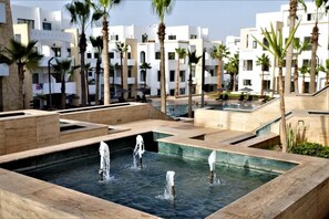 Un área de piscina con múltiples fuentes de agua y palmeras, rodeada de edificios modernos.