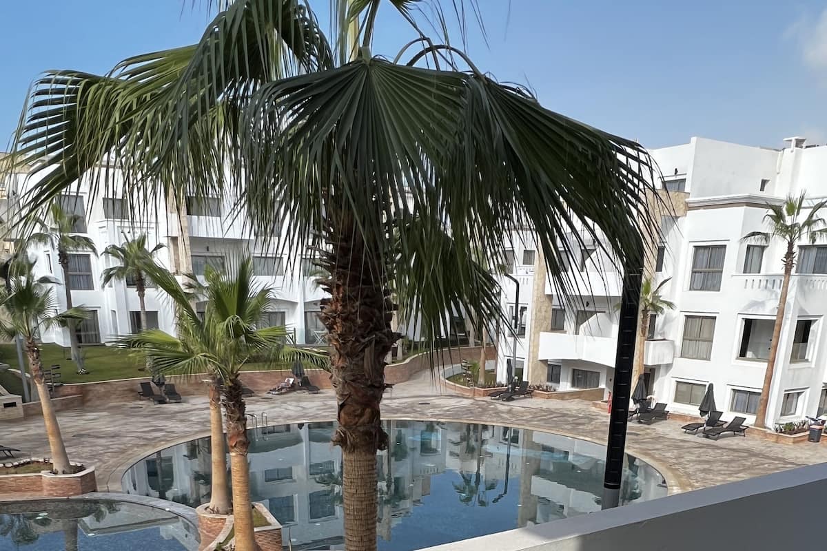 A balcony with wicker furniture, a pool, palm trees, and a modern building in the background.