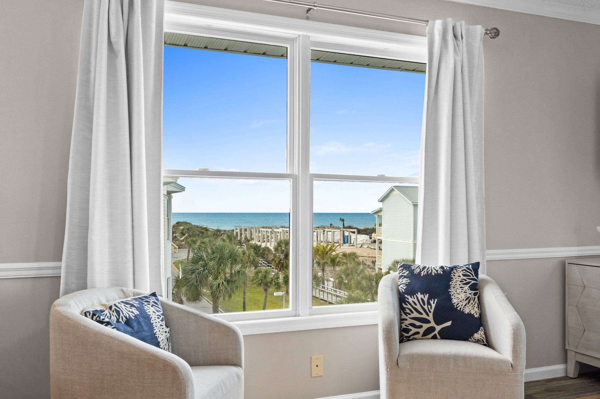 A room with a large window offering a view of the beach and ocean. There are two chairs with decorative pillows.