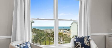 A room with a large window offering a view of the beach and ocean. There are two chairs with decorative pillows.