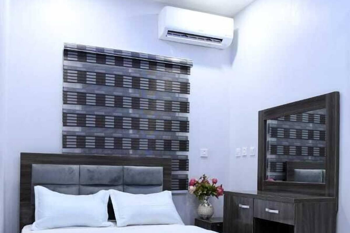 A modern bedroom with a large bed, a built-in air conditioning unit, a decorative wall panel, a mirror, and a vase with flowers.