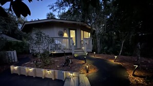 A cabin with a porch, wooden deck, and pathway leading to it, surrounded by trees. - Relaxing 2 BR cottage, walking distance to beach (Hilton Head Island)