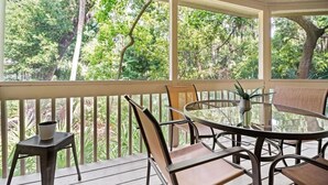 A covered outdoor seating area with a glass table and chairs, overlooking a forested area. - Relaxing 2 BR cottage, walking distance to beach (Hilton Head Island)