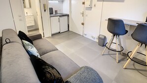 Een knus woongedeelte met een bank, een kleine keukenhoek, een aan de muur bevestigde televisie en een eethoek met twee stoelen.