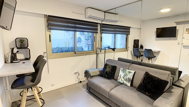 Een moderne woonkamer met een grijze bank, een klein bureau met stoel, een televisie en een raam met jaloezieën.
