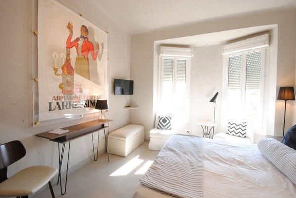 A bedroom with a bed, a desk, a chair, a television, and a window with shutters.