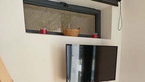 Een kamer met een flatscreen televisie aan de muur, een klein houten kastje met een afstandsbediening, en een raam met een mand en twee rode bekers op de vensterbank.