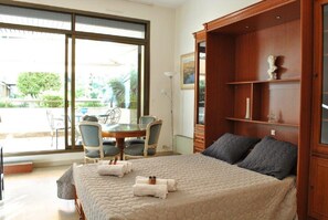 A bedroom with a large bed, a wooden wardrobe, a dining table with chairs, and a view through a large window.