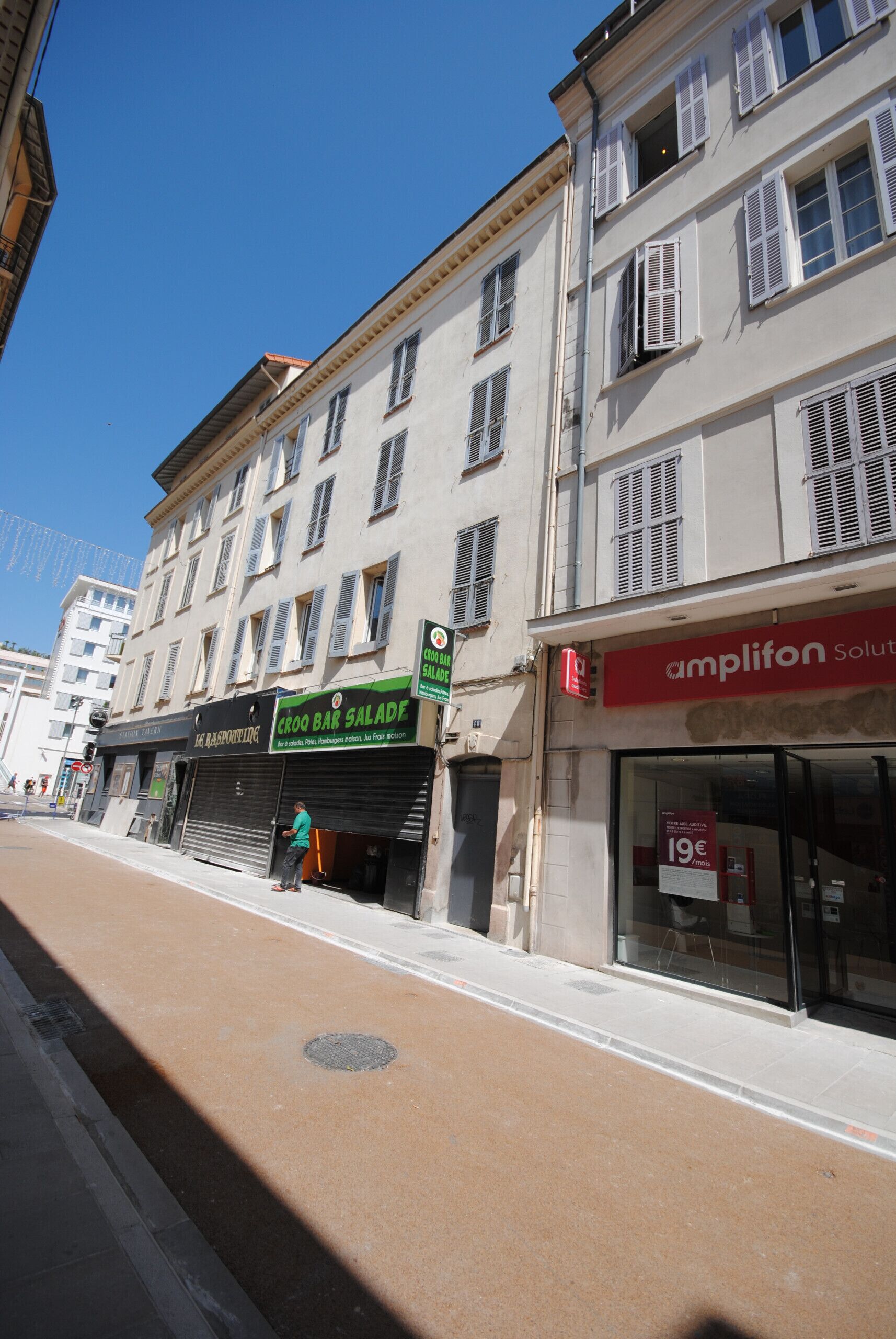 Une vue de rue avec des bâtiments, dont un magasin fermé reconnaissable à son enseigne verte et à son auvent rouge.
