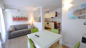 A modern living room with a sofa, a dining table with chairs, a kitchen area with cabinets, a painting on the wall, and a lamp. - Palais Rouaze/ Beg4012/ 2P (Cannes)