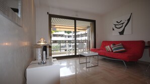 Un salon moderne avec un canapé rouge, une table basse blanche et une lampe. Une grande fenêtre offre une vue sur les bâtiments à l’extérieur.