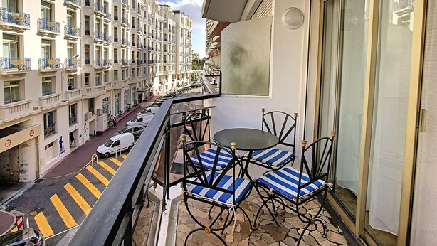 Un balcón con mesa y sillas, con vista a una calle con coches aparcados y una hilera de edificios con balcones.