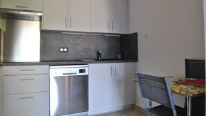 A kitchen with white cabinets, a stainless steel dishwasher, and a dining area with a chair and table. - WINDSOR/DET2002-1/bed (Cannes)