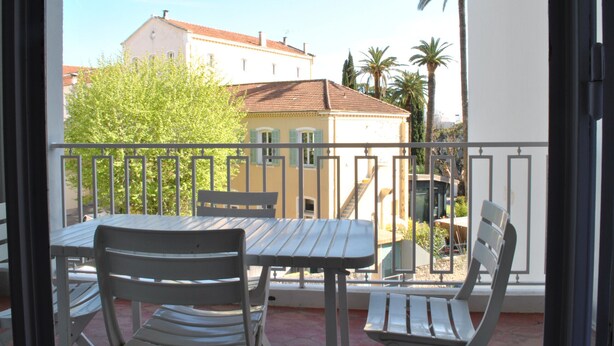 A balcony with a table and chairs, overlooking a building and palm trees. - WINDSOR/DET2002-1/bed (Cannes)